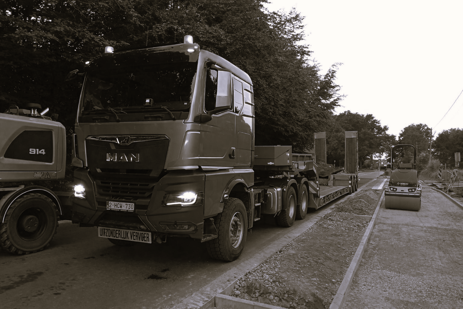 Grijze MAN vrachtwagen met oplegger op bouwweg, met gele wals en verkeersborden langs bomen.